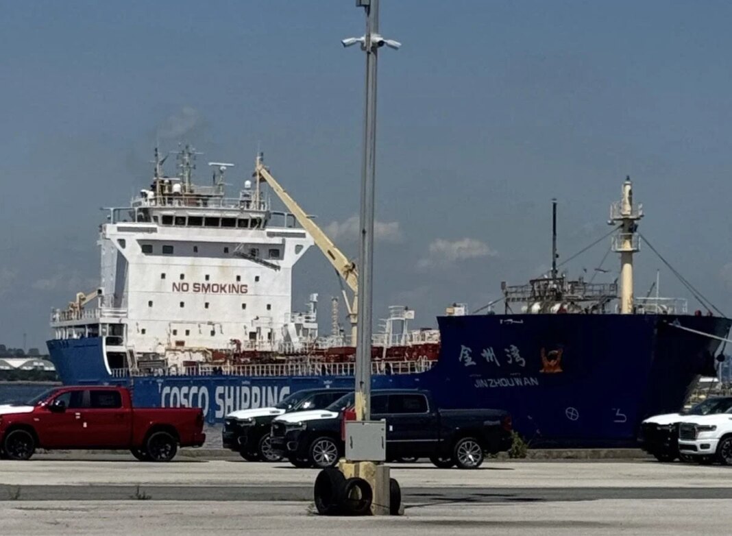 American Mariners Protest Chinese Tanker as Jones Act Waiver Faces Growing Scrutiny Chinese-flagged asphalt tanker JIN ZHOU WAN berthed in Baltimore after transporting asphalt from New Jersey under the emergency Jones Act waiver. The blue COSCO Shipping vessel is docked behind parked vehicles along the waterfront.