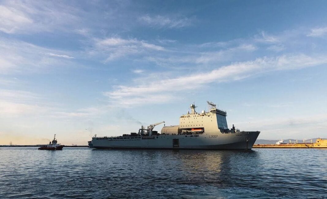 Hormuz Strikes Threaten Progress on Europe’s De-mining Mission RFA Lyme Bay sails from Gibraltar escorted by a tugboat during a previous deployment, with calm water and blue skies surrounding the Bay-class support ship.